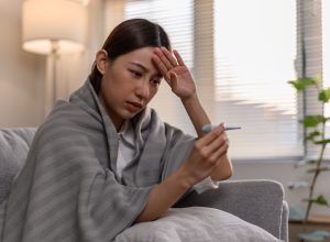 A sick young woman wrapped in a blanket on her couch, taking her temperature