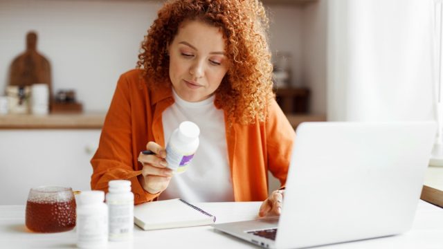 Redhead nutritionist working from home on laptop at her article about deficiency of food supplements and its influence on human body and health sitting at kitchen table with vitamin bottle