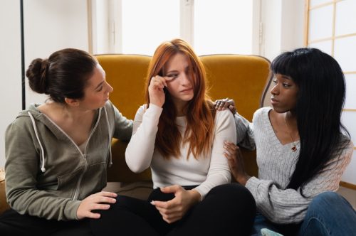 close-up three friends in a living room. Woman in the middle is crying and the other two females are giving comfort and support.