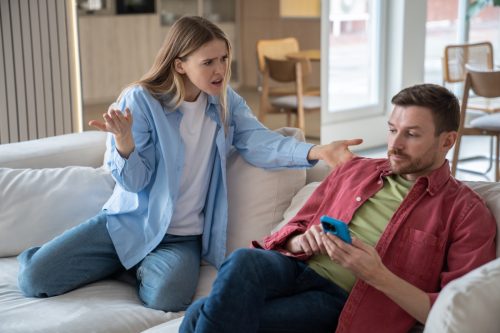 Indignant, offended, hysterical woman screams at husband sitting on sofa with cellphone. Couple family quarrel conflict misunderstanding discord. Indifferent man ignores dissatisfied wife's questions.