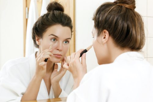 Woman in white robe looking in the mirror, using concealer for under eye circles
