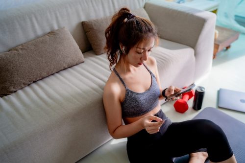 Woman in workout clothes sitting on living room floor ordering something on her phone