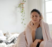 woman sitting up in bed with a blanket wrapped around her, holding her throat to indicate a sore throat