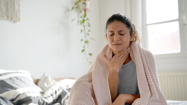 woman sitting up in bed with a blanket wrapped around her, holding her throat to indicate a sore throat