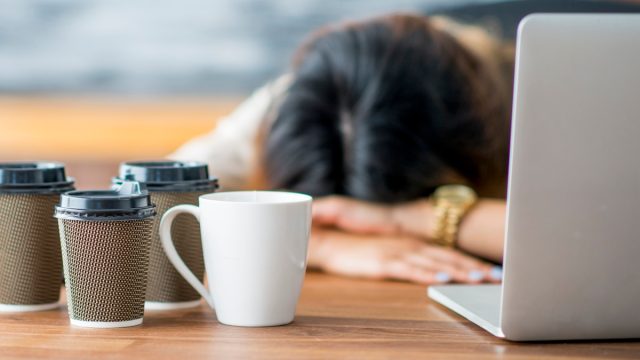 Woman asleep at her desk behind her laptop with several coffee cups in front of her