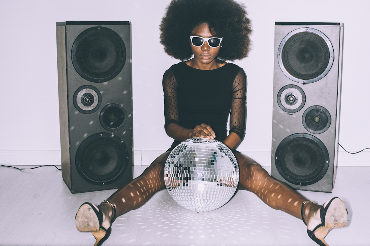 woman in black dress and sunglasses sitting on floor and holding disco ball next to two large speakers