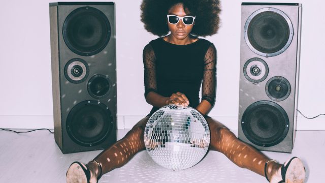 woman in black dress and sunglasses sitting on floor and holding disco ball next to two large speakers