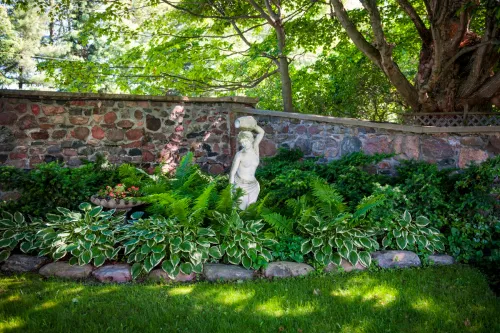 Lush green summer garden with perennial plants and statue near stone wall