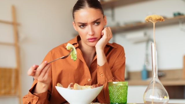 upset-looking woman sitting alone at table eating caesar salad