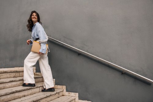 In full growth, young caucasian woman with documents in hands climbs stairs going to work. Brunette wears blue shirt, white trousers and sweatshirt. Business lady concept