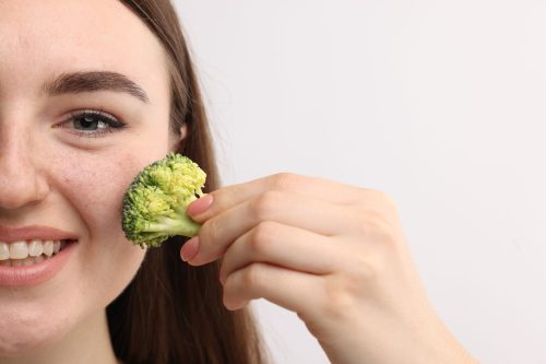 Smiling woman making fake freckles with broccoli and cosmetic product on light background, closeup.