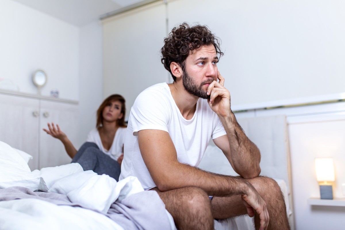 upset man sitting on edge of bed while a woman is upset in the background