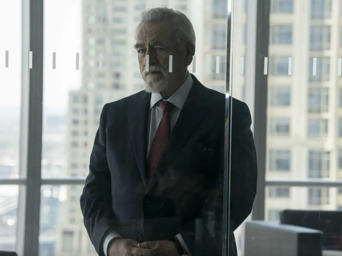 Actor Brian Cox playing Logan Roy in Succession. He is standing behind a glass door in his office.