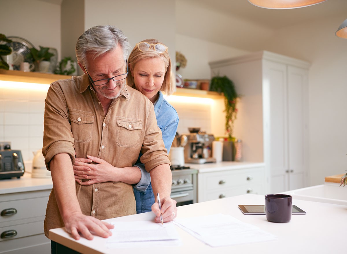 A woman wraps her arms around her partner's waist as he writes on paper on the counter.