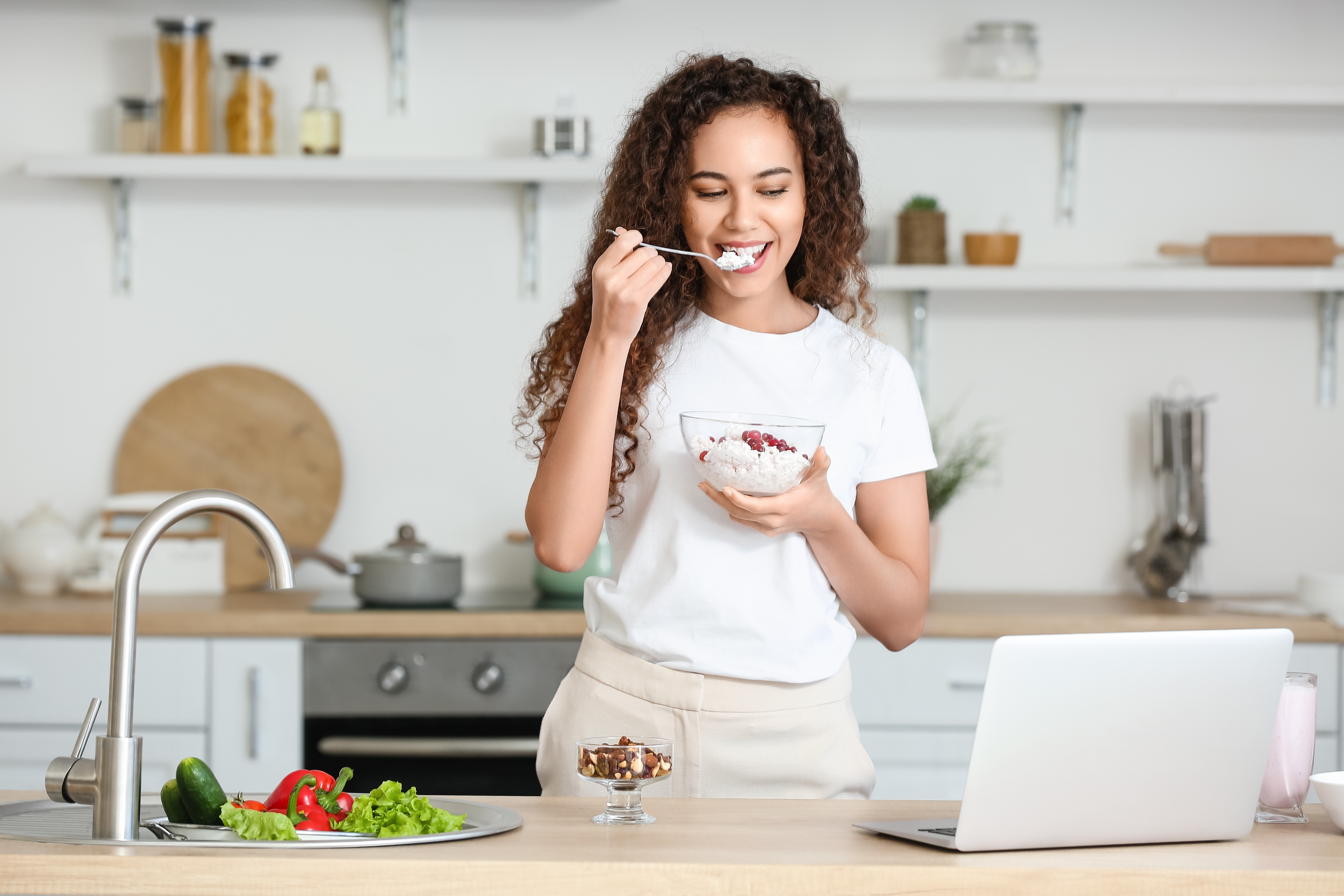 A woman eats cottage cheese, one of the best snacks for weight loss, from a bowl in a sunlit kitchen with white walls.