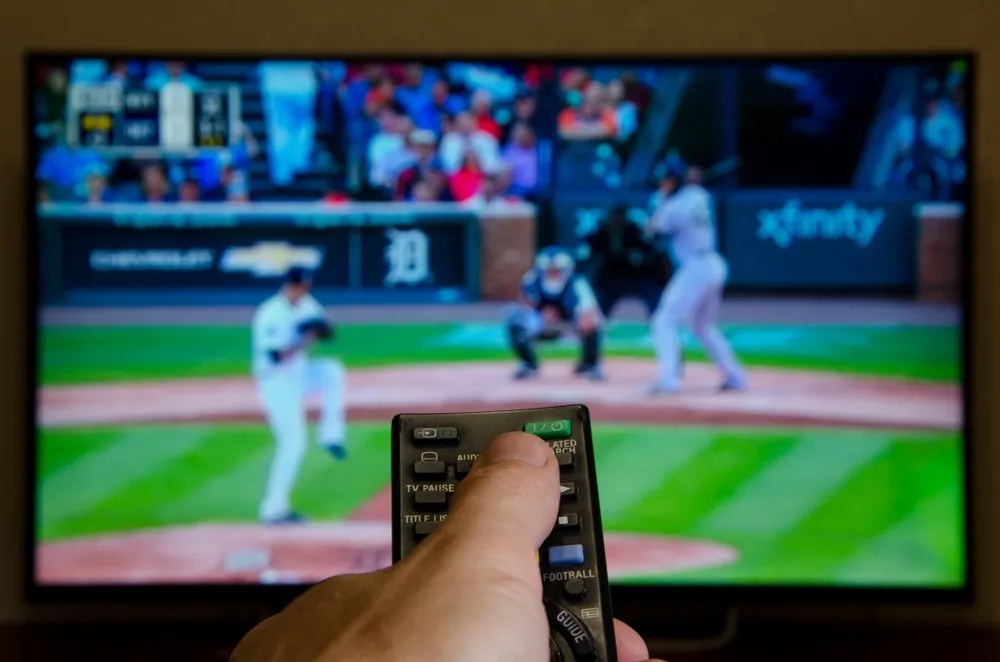 Closeup of a person holding a remote aimed at a TV showing a baseball game