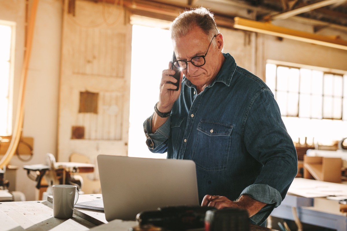 Mature carpenter working on laptop and answering phone call in his carpentry workshop.