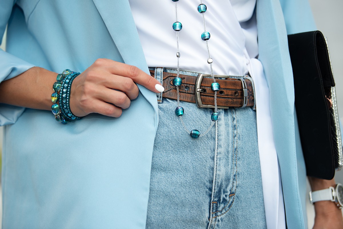 Detail of the body of a model dressed in light blue high-waisted jeans, an ornamented leather belt and a blue jacket.