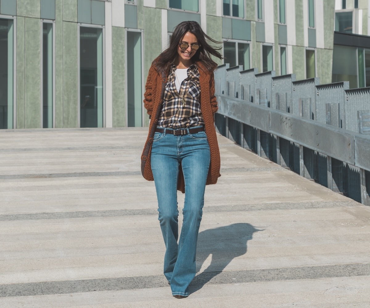 stylish woman wearing plaid shirt, long sweater, and flare jeans walking down a ramp in a city