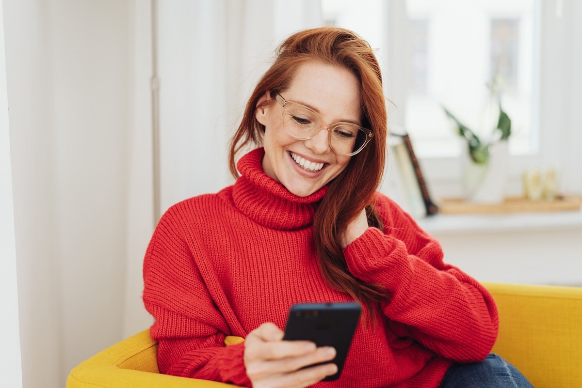 woman with long red hair wearing a red sweater, sitting on a yellow chair, using her phone