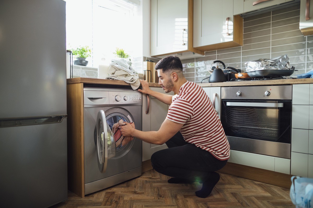 Man crouching down to put washing into his washing machine in his kitchen