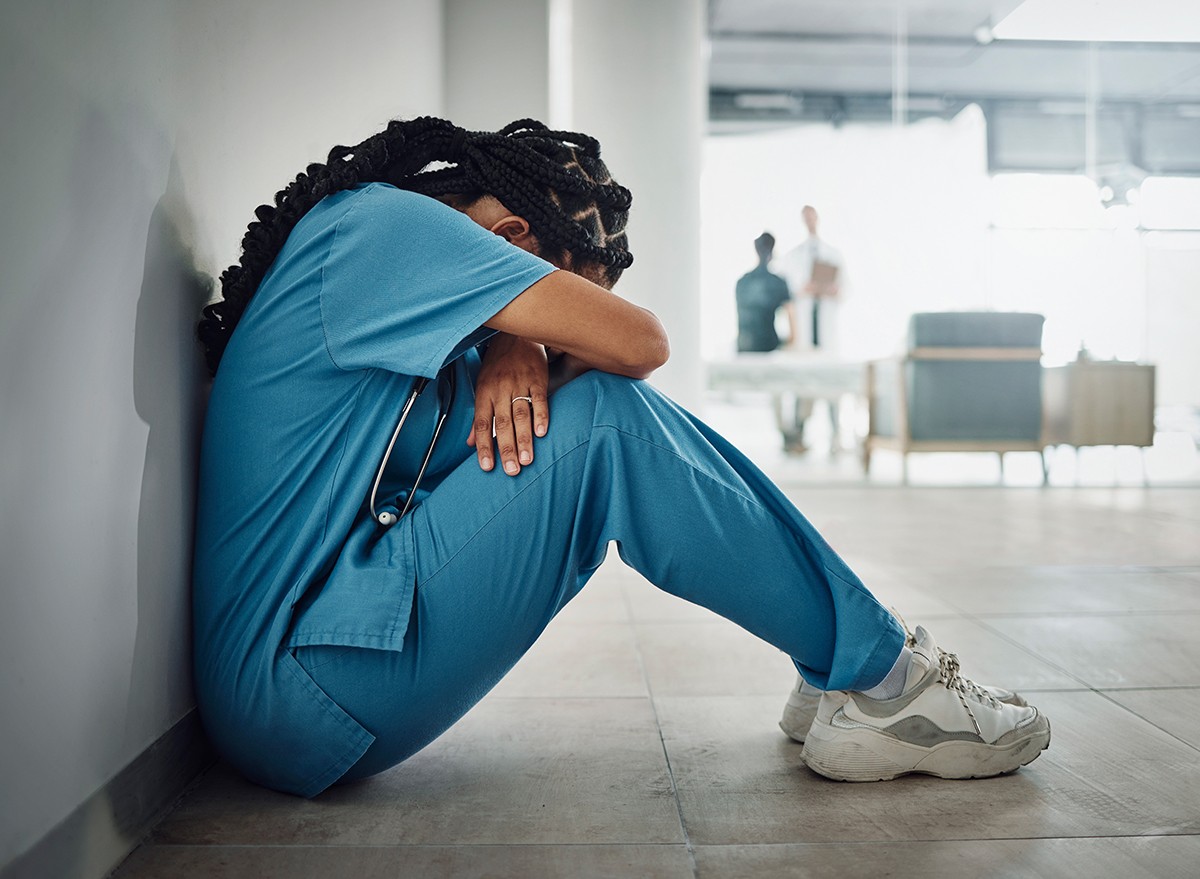 A nurse sits alone in a hallway looking upset