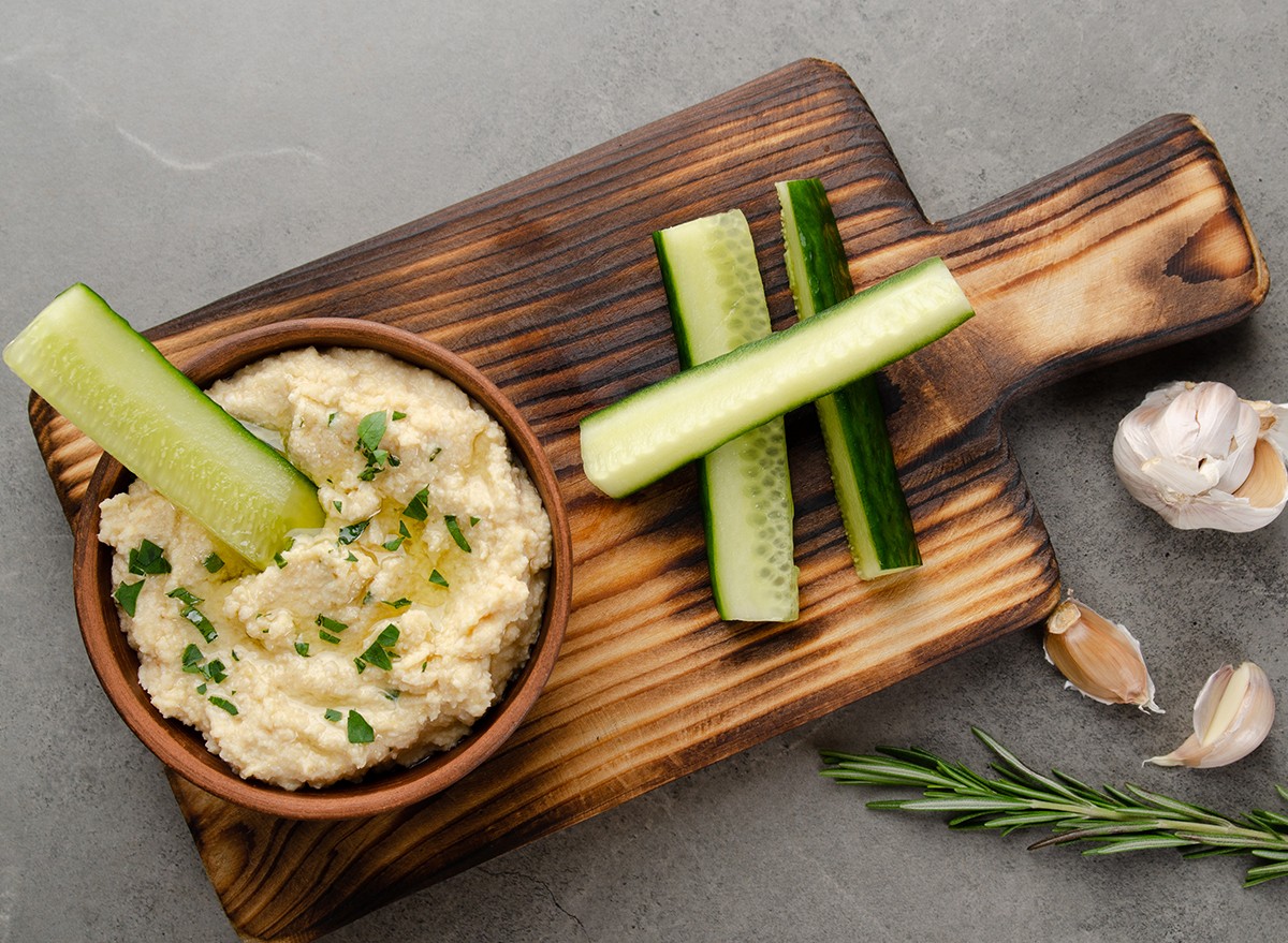 Cut up cucumbers and a bowl of hummus on a wooden board