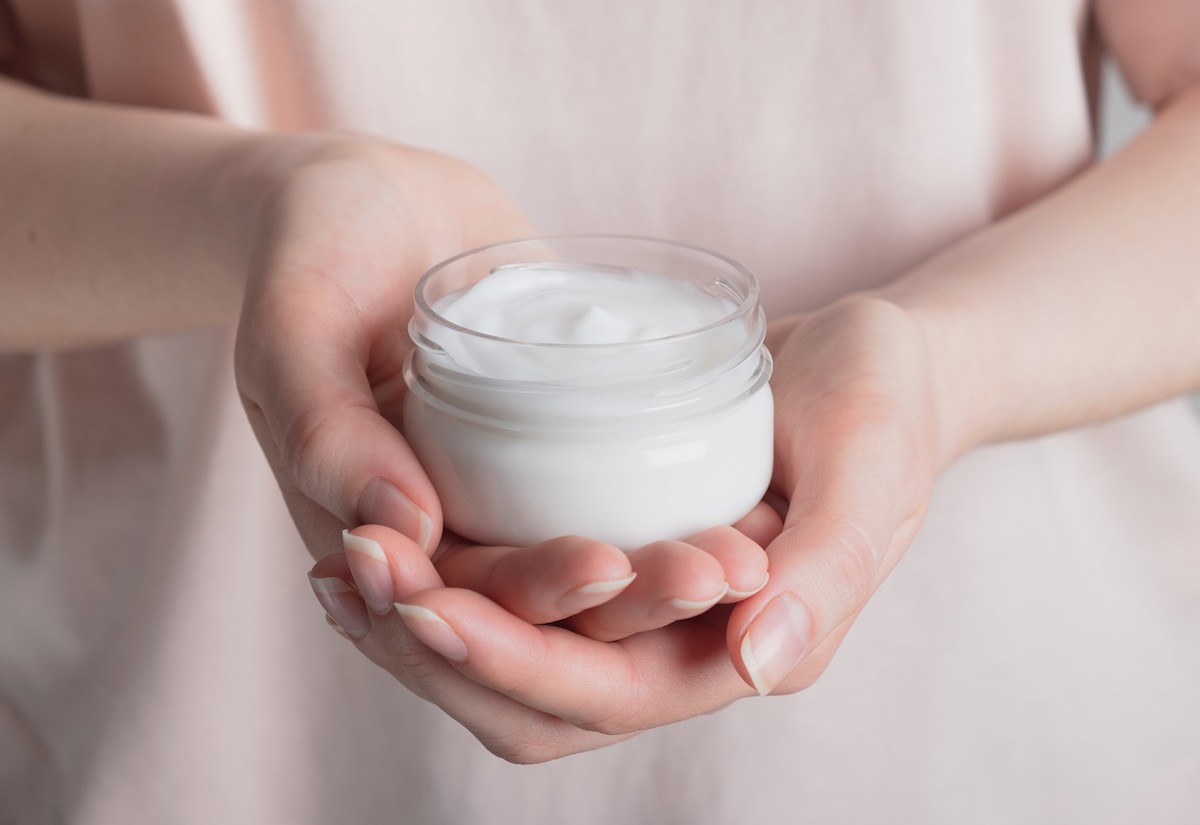 Closeup of a woman in a pale pink shirt holding out a jar of face cream