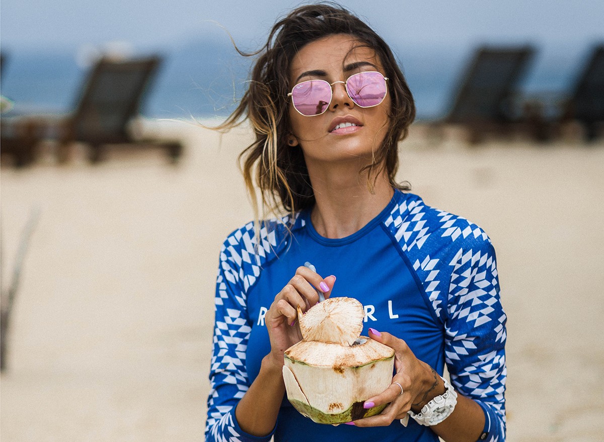 Woman on the beach wearing an SPF protection sun shirt, drinking coconut water