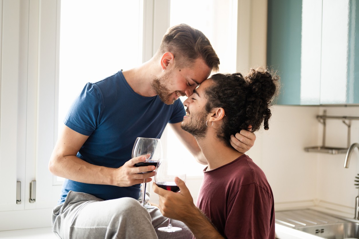 male couple having fun and drinking red wine in their kitchen while cuddling
