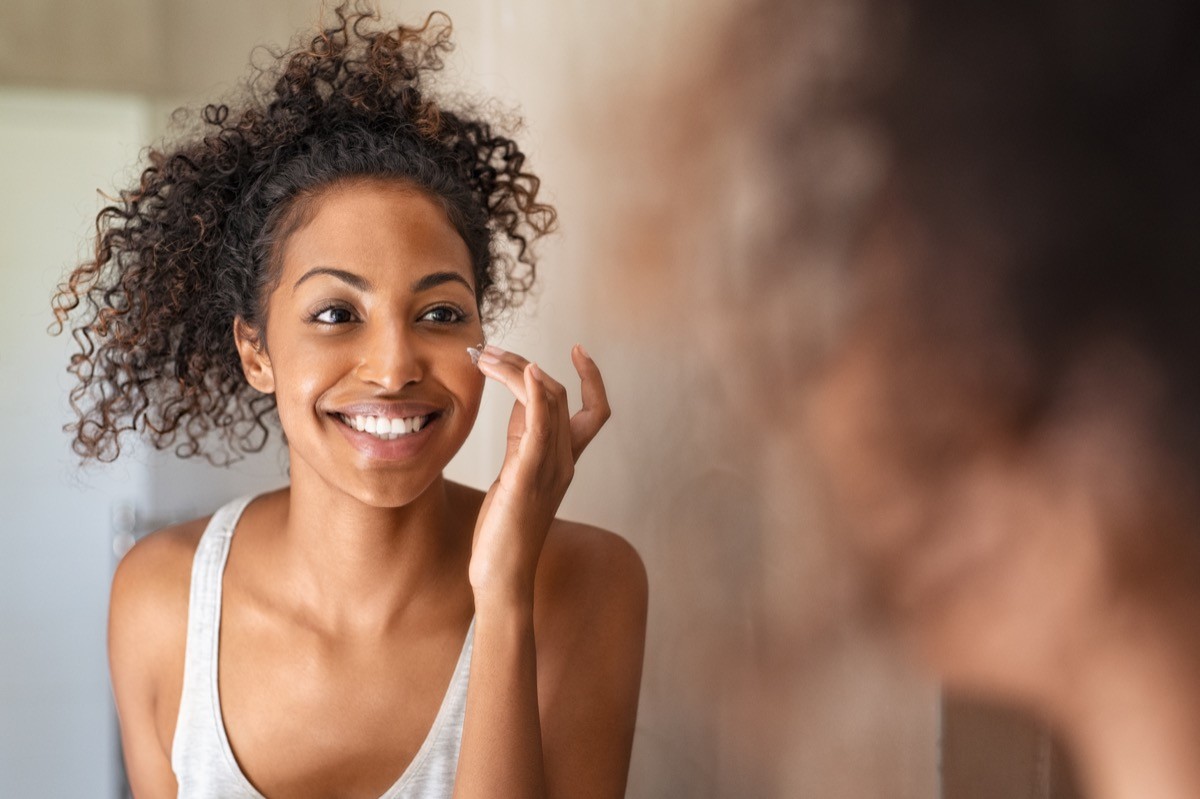 A woman applying moisturizing cream to her face while standing in front of the mirror in bathroom