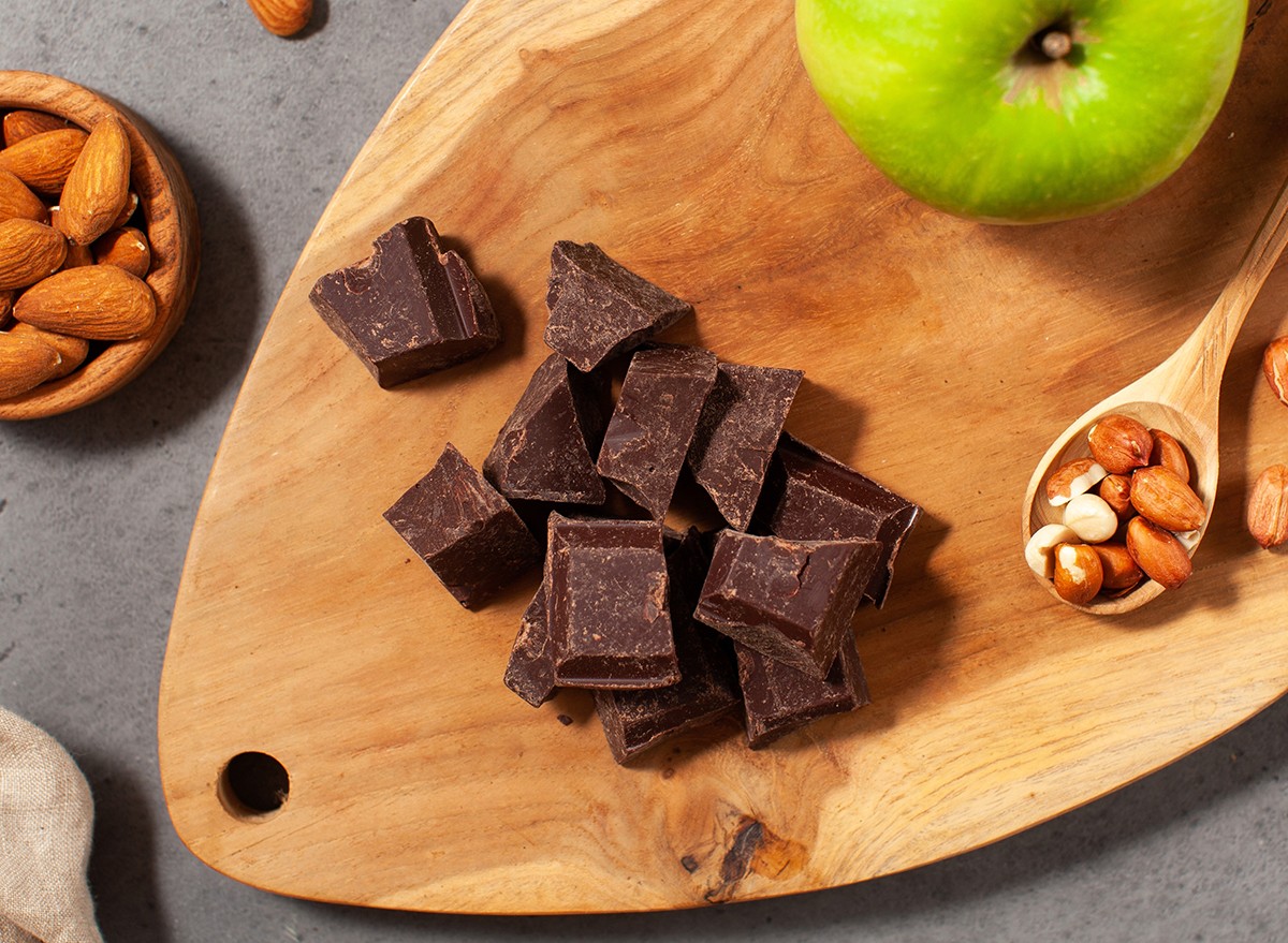A green apple on a wooden cutting board with pieces of dark chocolate and nuts