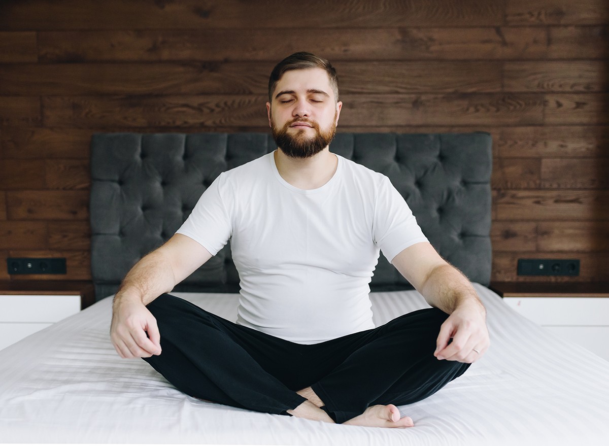 A man sitting cross-legged on a bed with his hands on his knees, eyes closed, doing breathing exercises