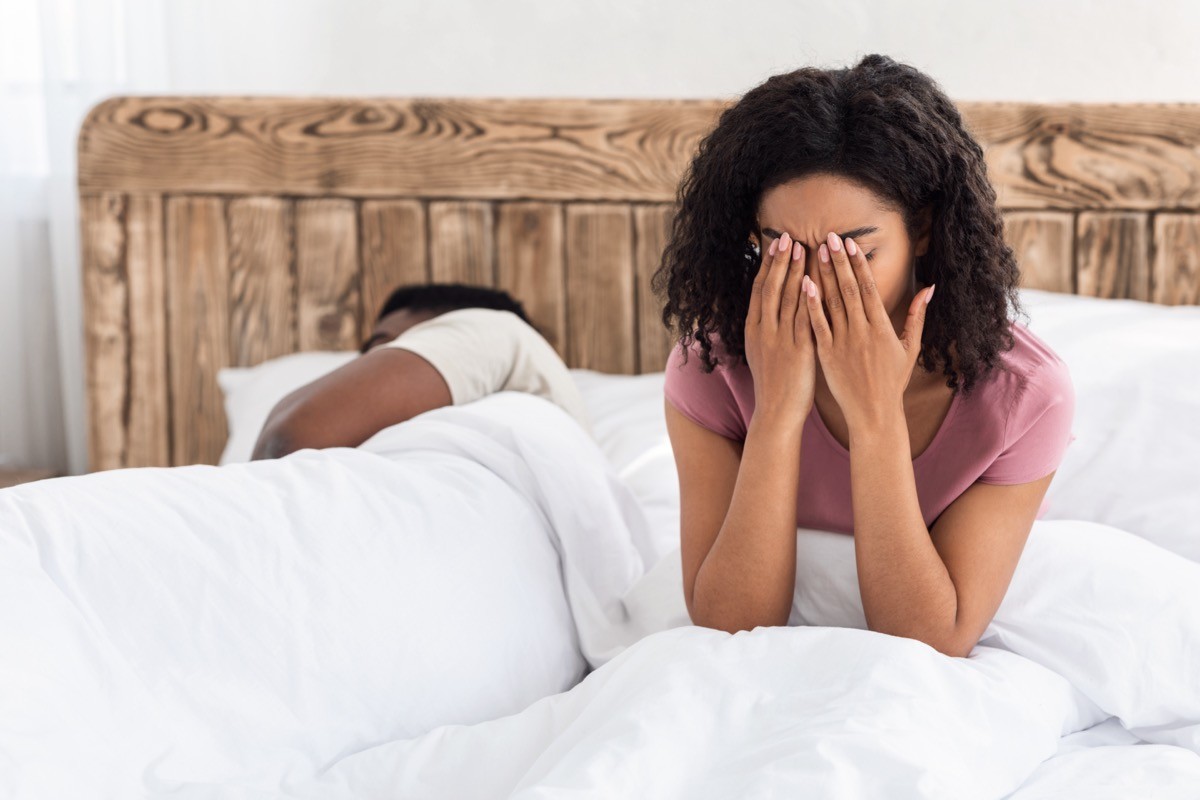 woman sitting up in bed rubbing her eyes while male partner sleeps
