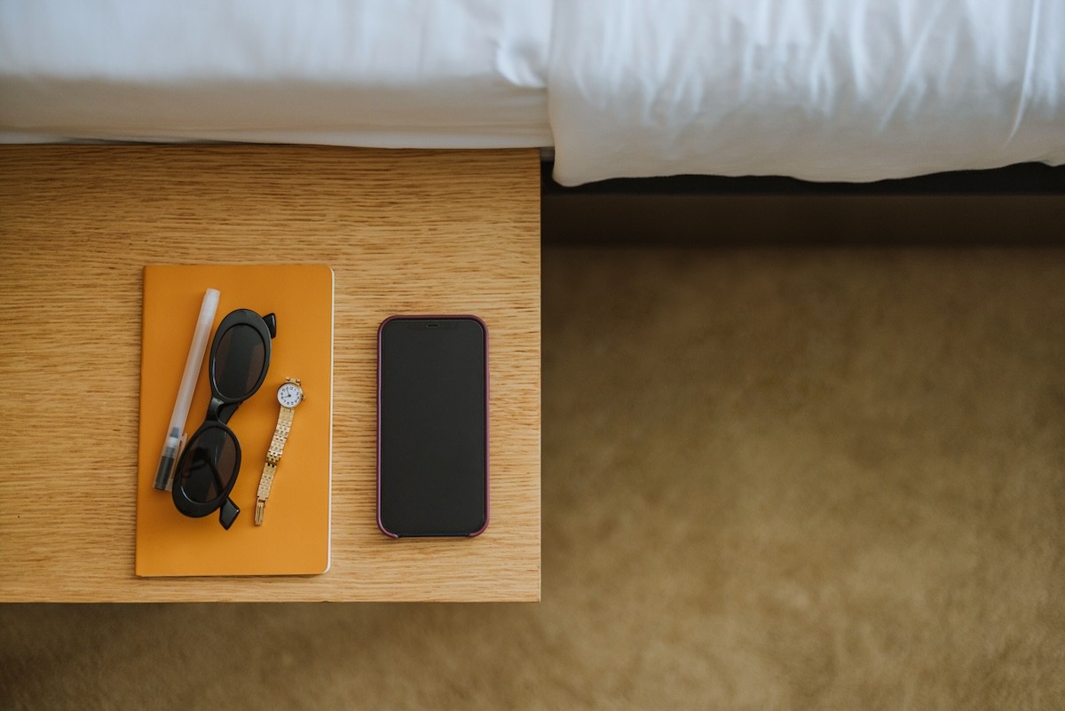 orange notebook, pen, sunglasses, watch, and phone on a nightstand next to bed
