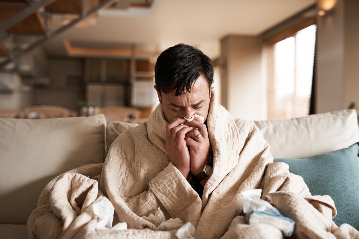 A man wrapped in a blanket sits on the couch blowing his nose with flu symptoms