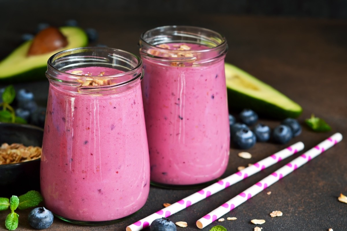 Two glasses of purple smoothies surrounded by blueberries and avocados