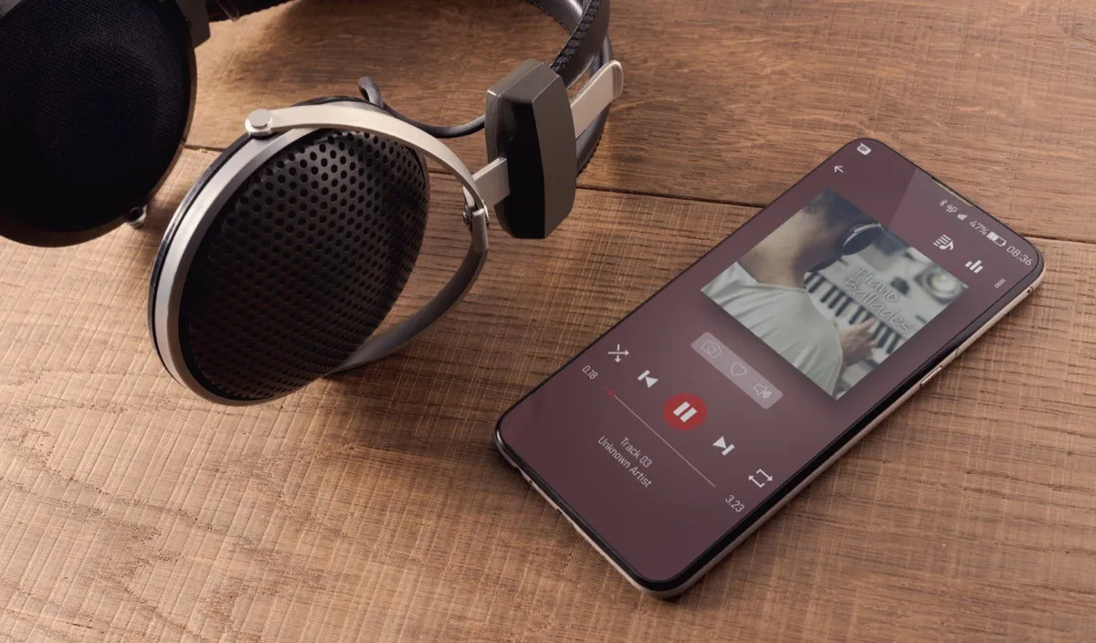 A pair of headphones and a smartphone on a wooden table. On the display you see the player, and a music album playing. Concept of new ways to relax anywhere.