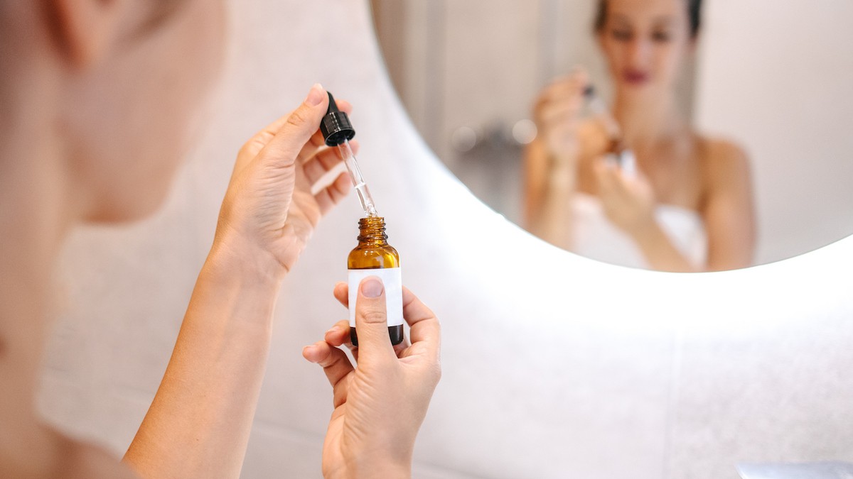 Woman in front of a mirror holding face oil for daily skincare routine in a bottle with a dropper