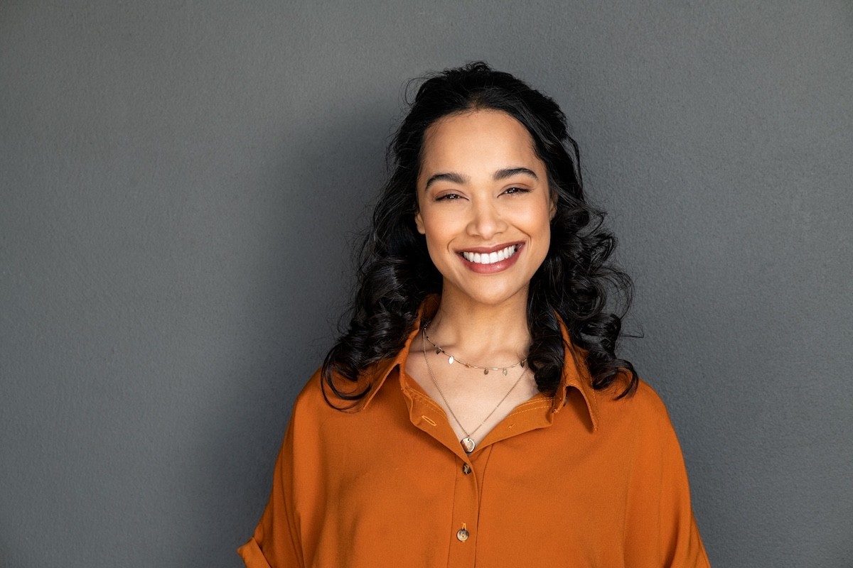 woman with dark hair wearing a burnt orange blouse standing against a gray wall