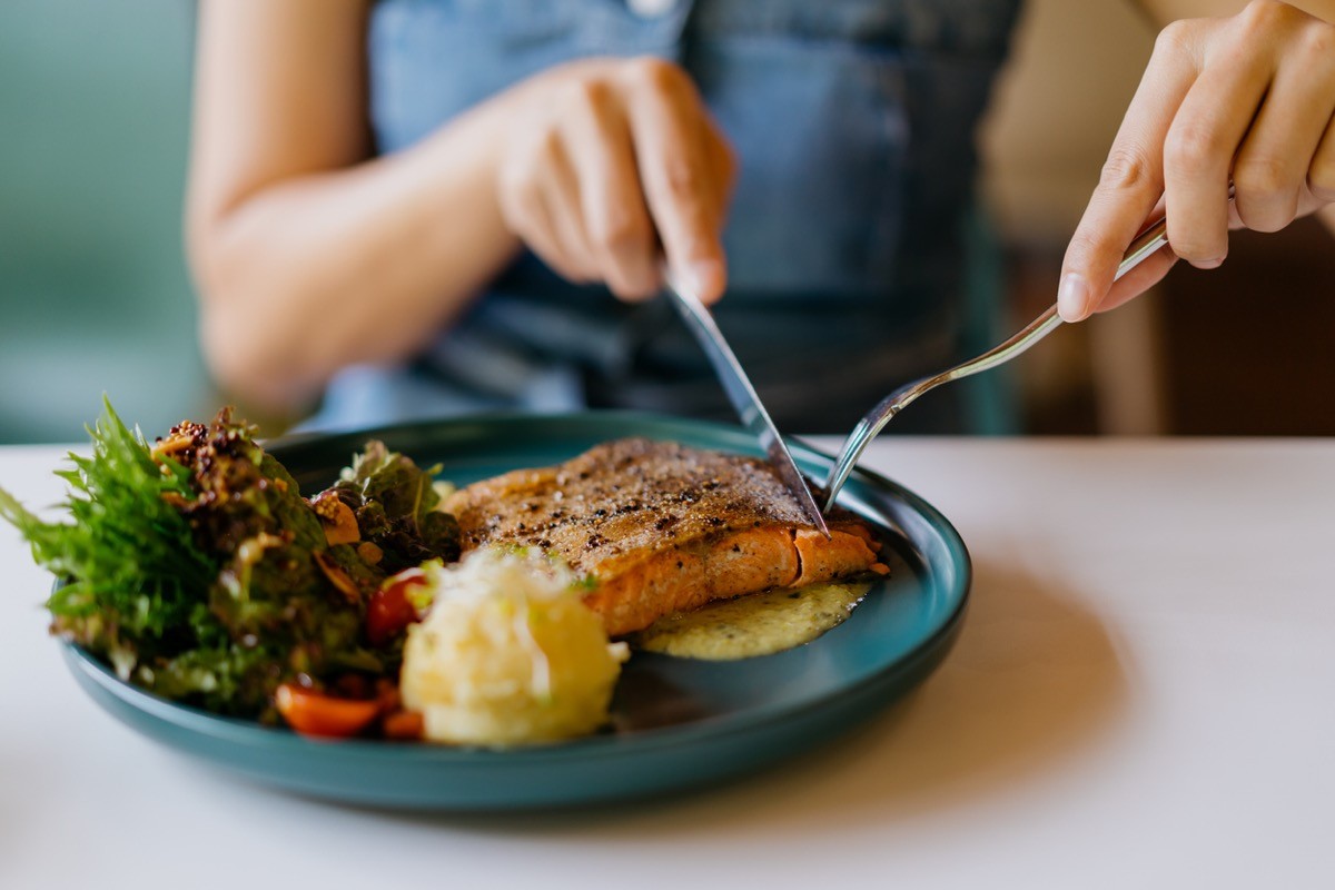 Close up shot of woman eating pan fried salmon with table knife and fork in cafe