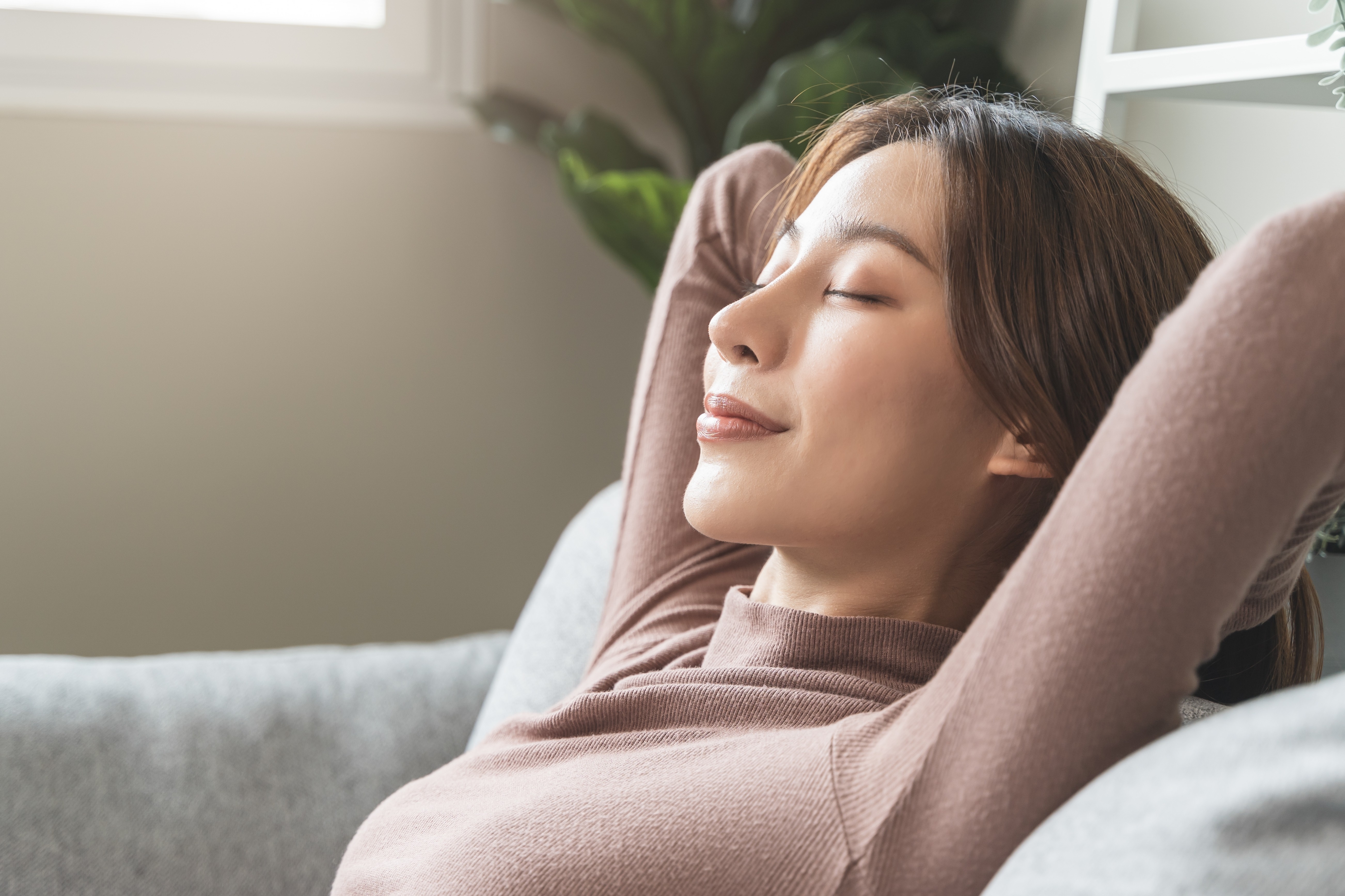 Close up of a calm-looking young woman on the couch with her hands behind her head.