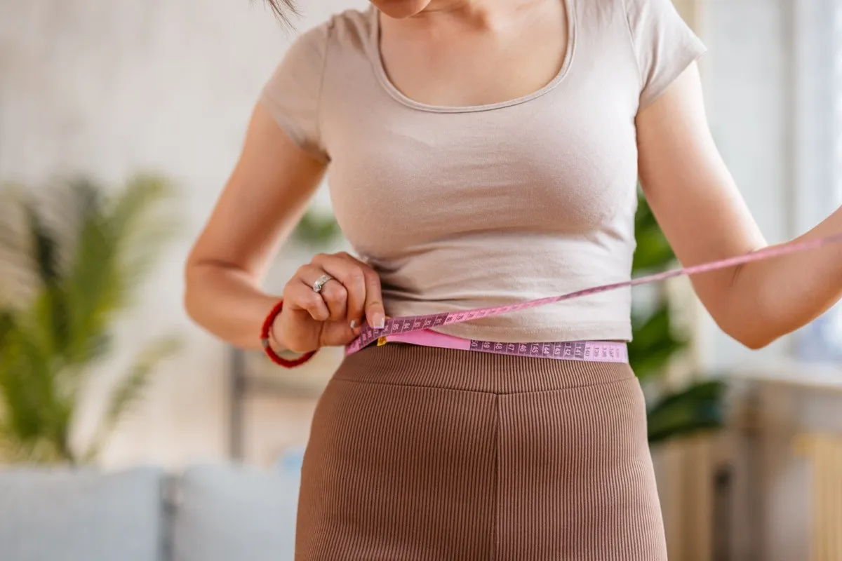 woman measuring her waist with a measuring tape in the living room at home.