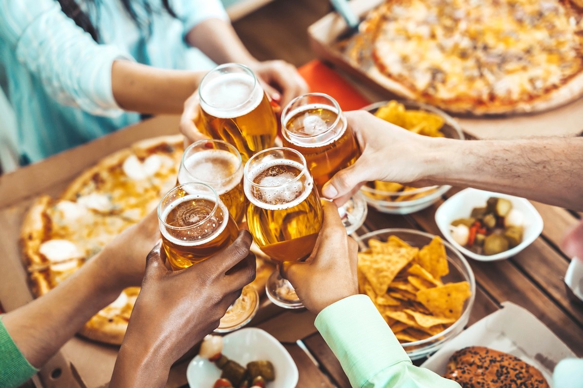 overhead view of people having a toast with beer over a table with pizza