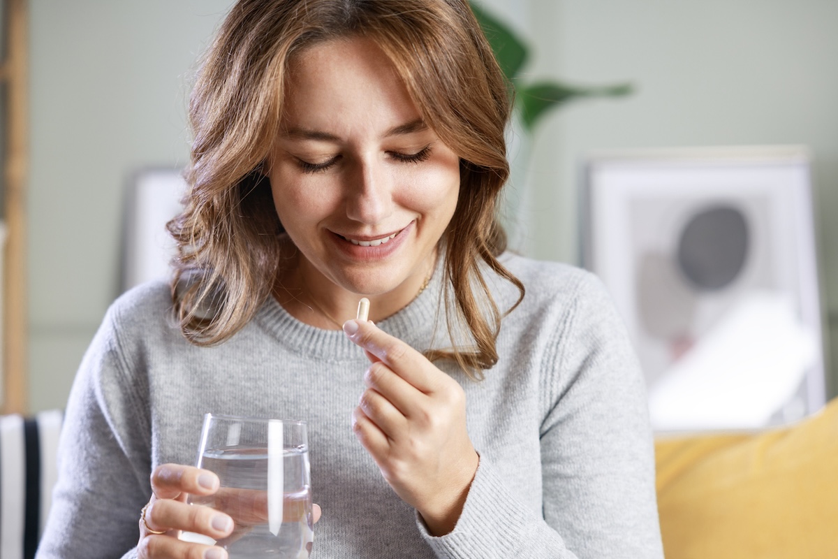 woman in a gray sweater sitting on her couch taking a supplement with a glass of water