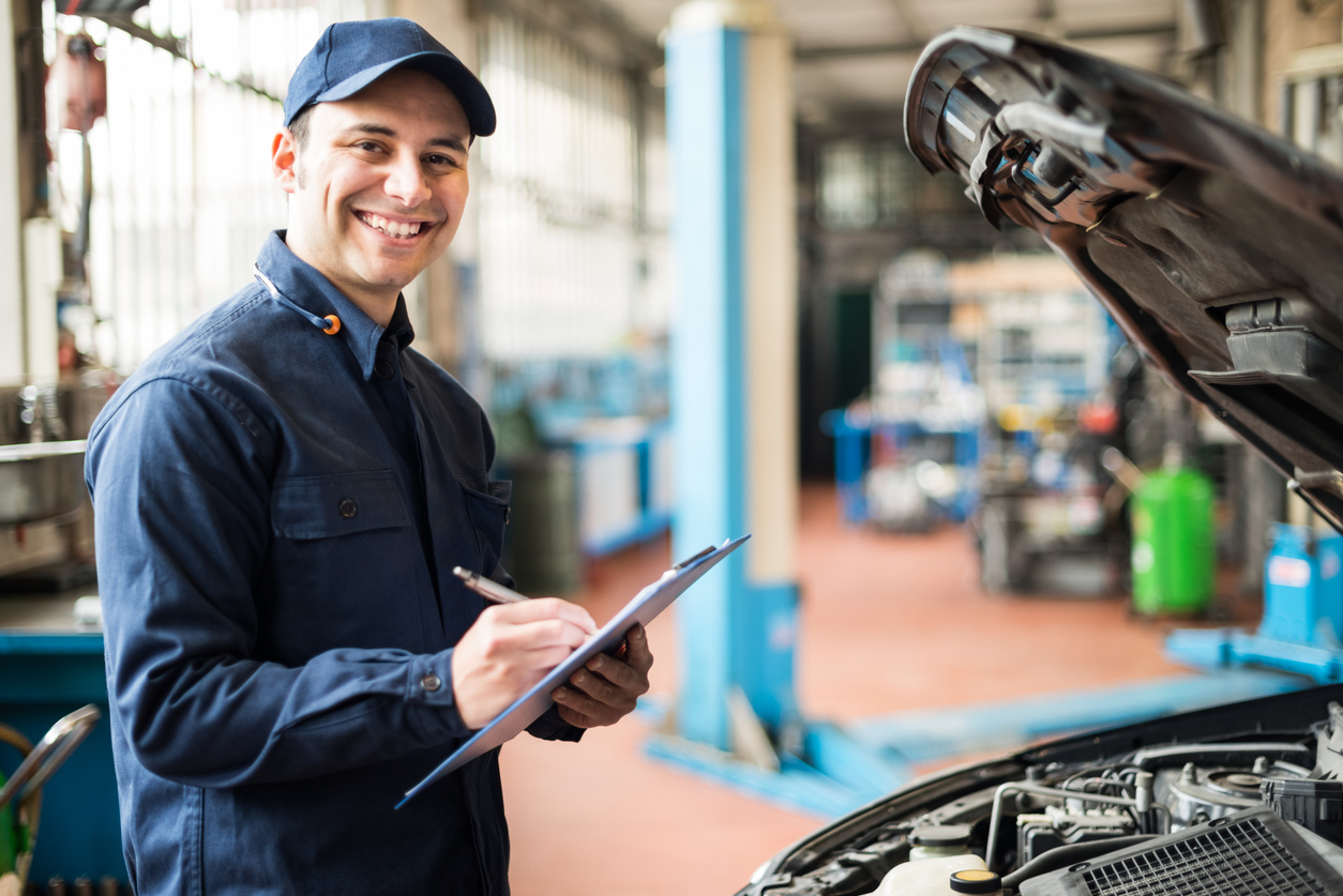 A mechanic smiling while holding a clip board and looking at a car with the hood up