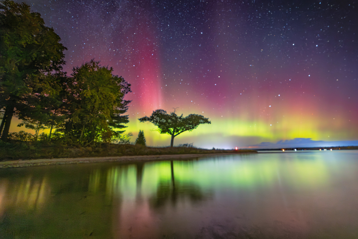 A vibrant aurora borealis lights up the night sky with shades of green, red, and purple, reflecting beautifully on a calm lake. Silhouetted trees on the shore add to the serene atmosphere under a starry sky.
