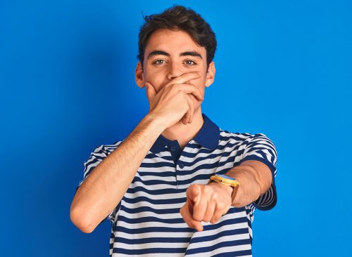 Teenager boy wearing casual t-shirt standing over blue isolated background laughing at you, pointing finger to the camera with hand over mouth, shame expression