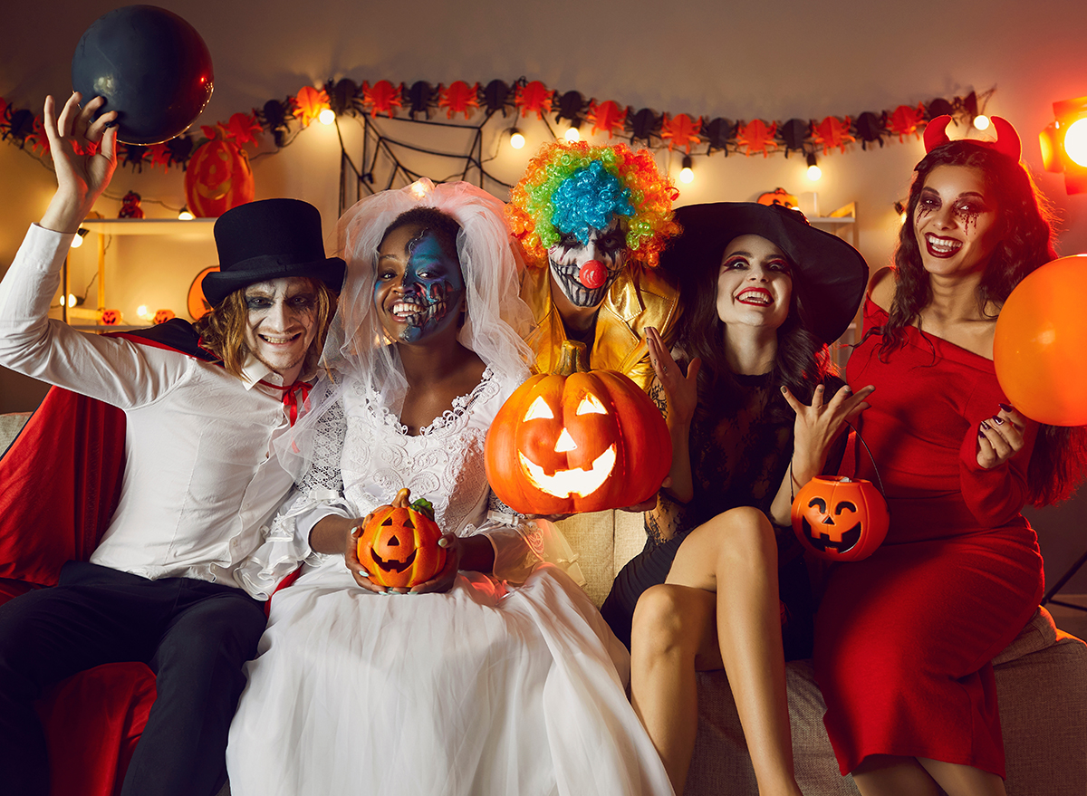 Portrait of group of cheerful crazy friends in various spooky costumes at fun Halloween party at home. Adult multiracial people sit on sofa with pumpkins and balloons in dark room and smile at camera.