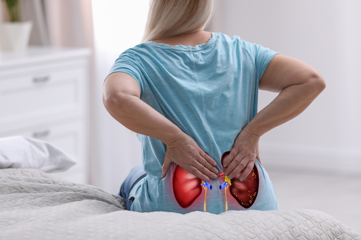 rear view of a woman in a blue t-shirt sitting on her bed clutching her lower back in pain, with a graphic of kidneys overlaid